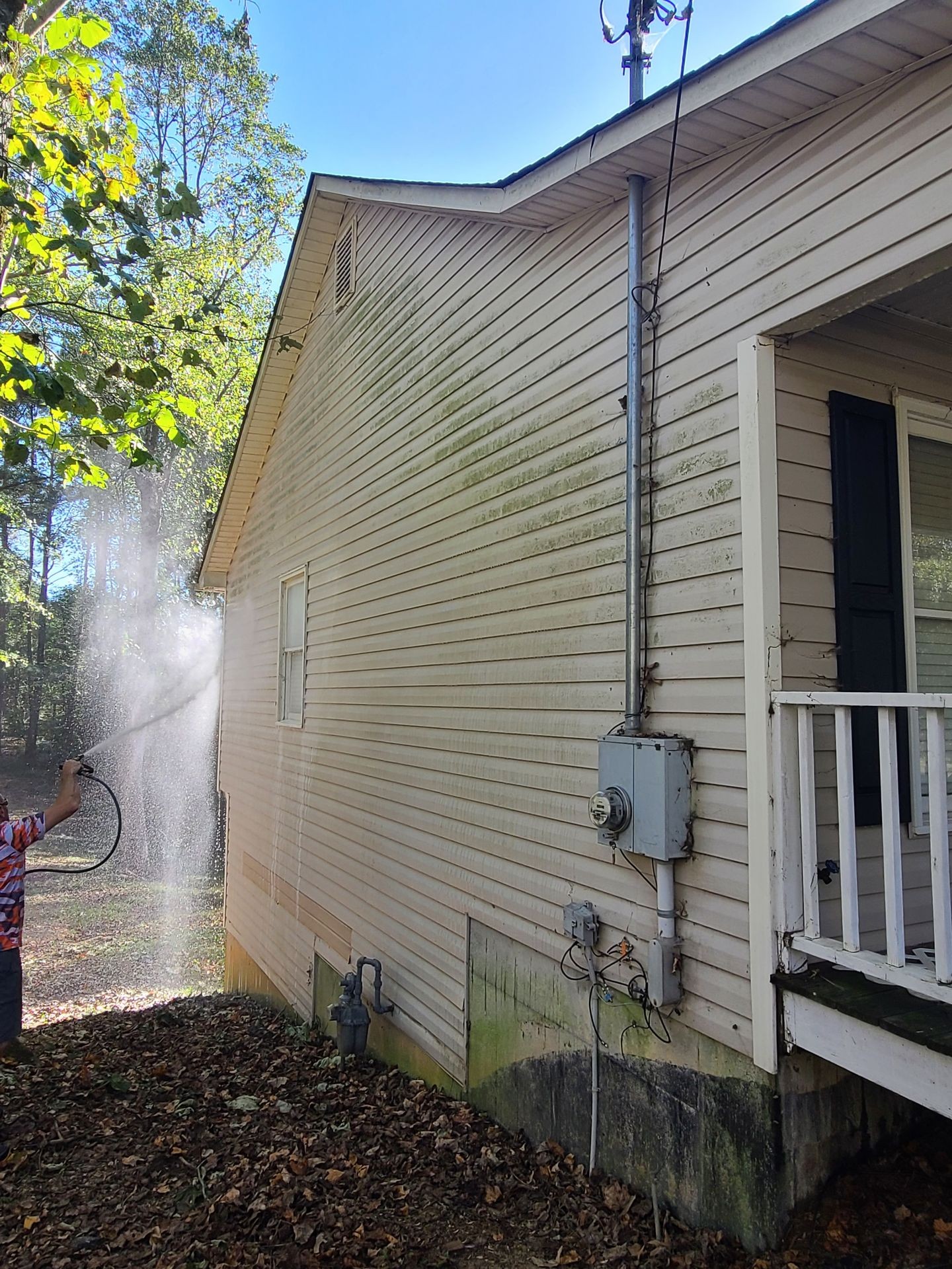 Person pressure washing the side of a house with vinyl siding stained by dirt and mildew.